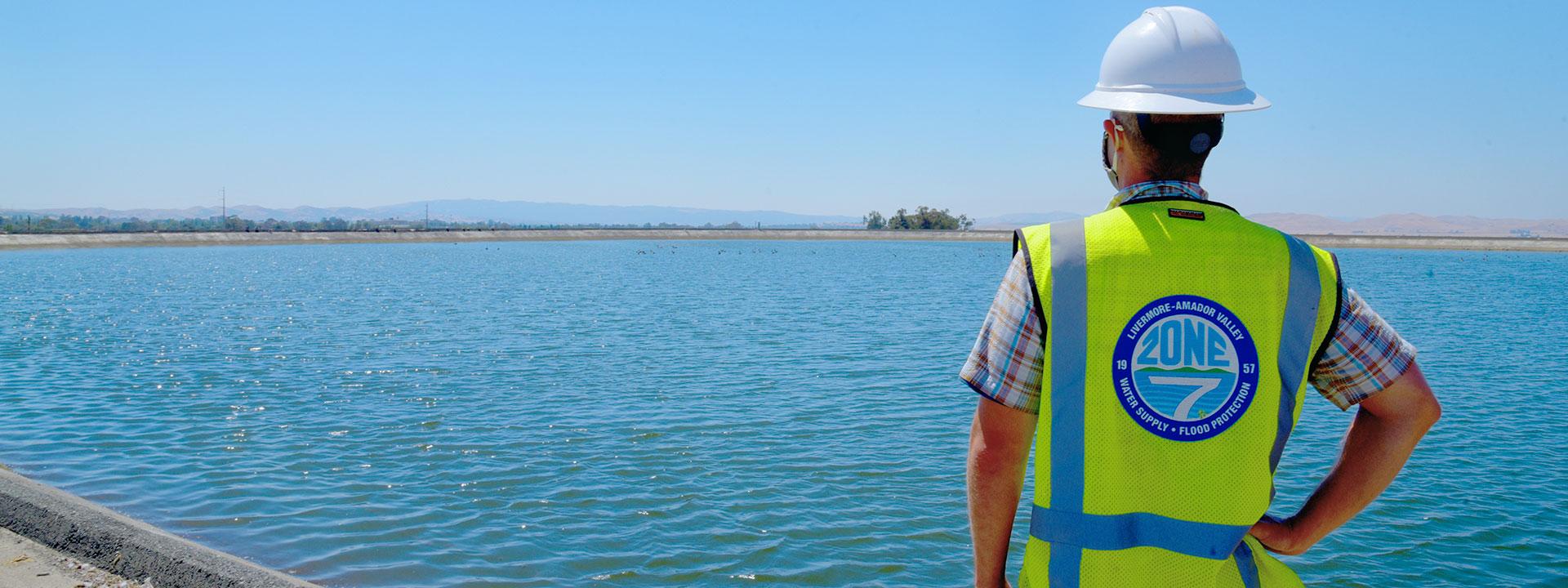 Zone 7 Engineer wearing a white hard hat and a yellow hazard vest looking out to a body of water