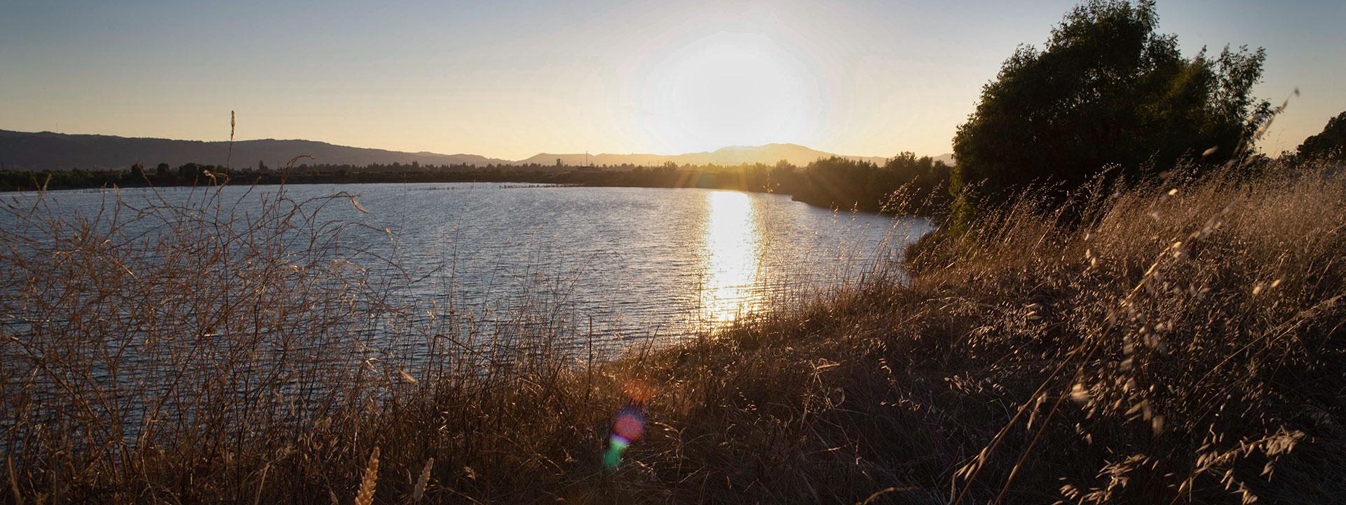 A blue lake surrounded by greenery and trees with a sunset reflected in the lake