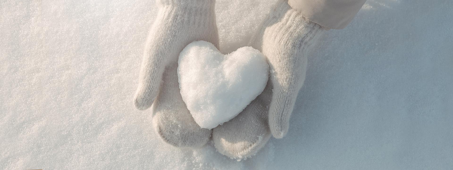 A child wearing white mittens delicately holding a heart-shaped snowball against a snowy background