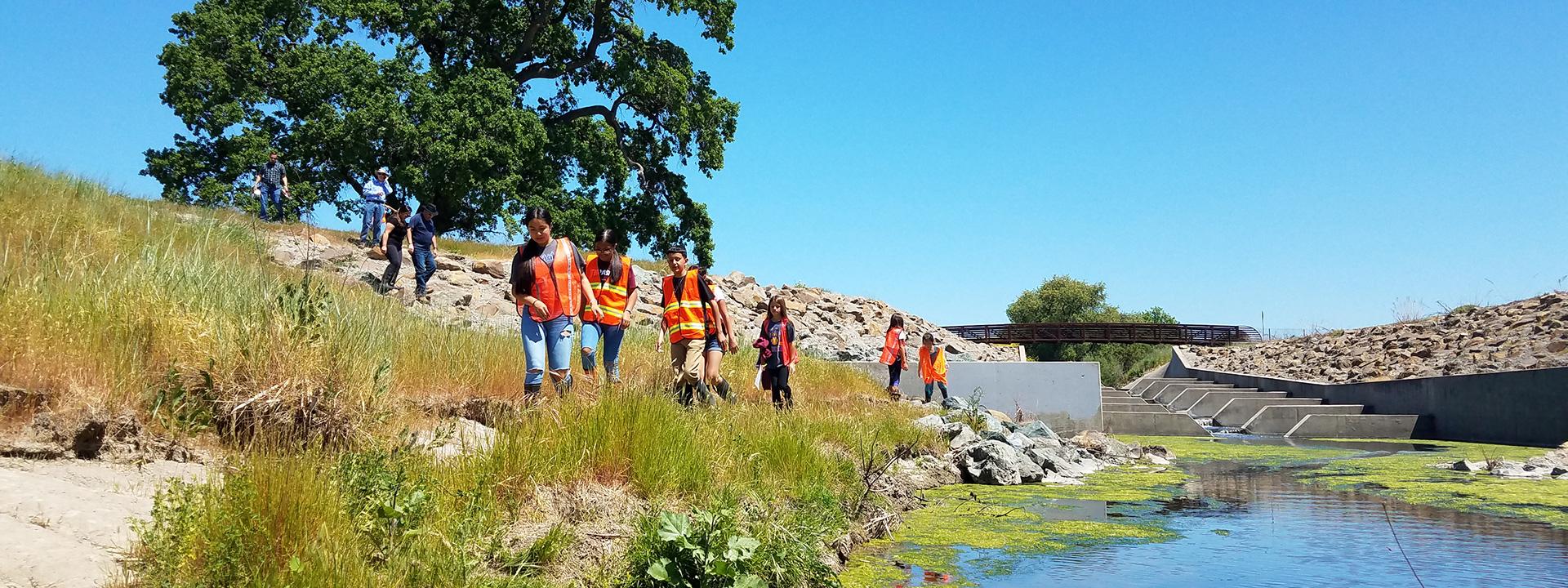 Children wearing orange safety vests walking along a creek