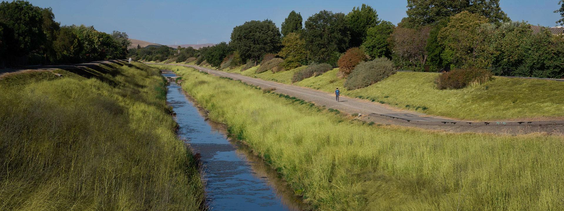 A blue stream alongside a trail in between two grassy banks