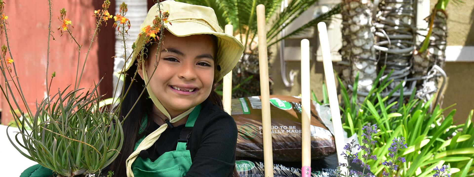 Child smiling wearing sunhat and gloves holding potted plant