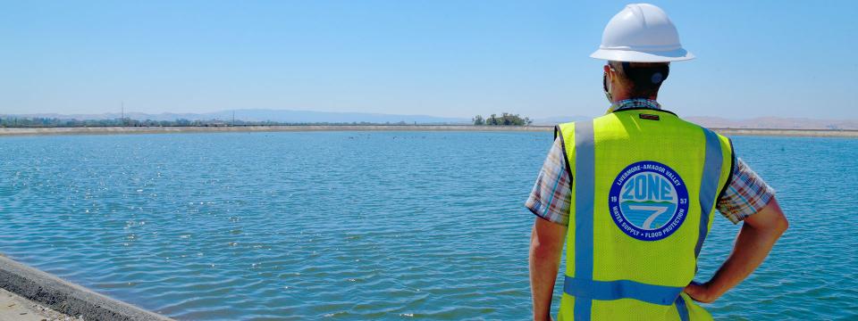 Zone 7 Engineer wearing a white hard hat and a yellow hazard vest looking out to a body of water