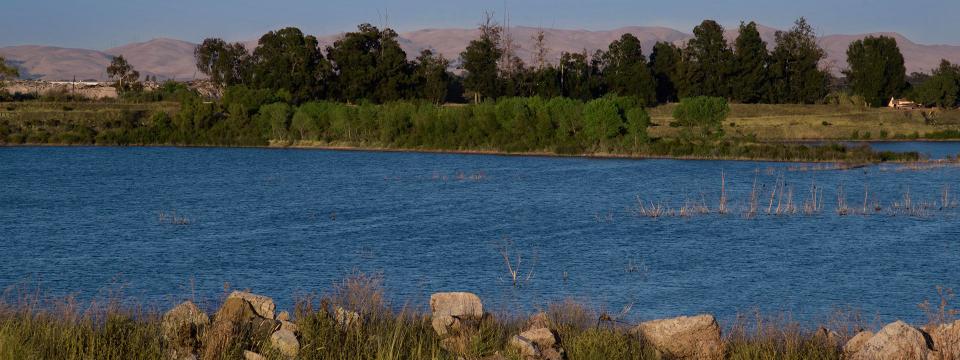 A large blue body of water surrounded by green brush and rocks with mountains in the background