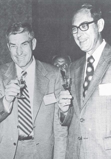 Two men smiling, wearing suits and nametags while raising drink glasses