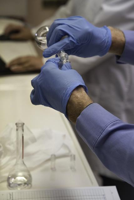 Lab technician with blue latex gloves pouring substance from a beaker into a test tube.