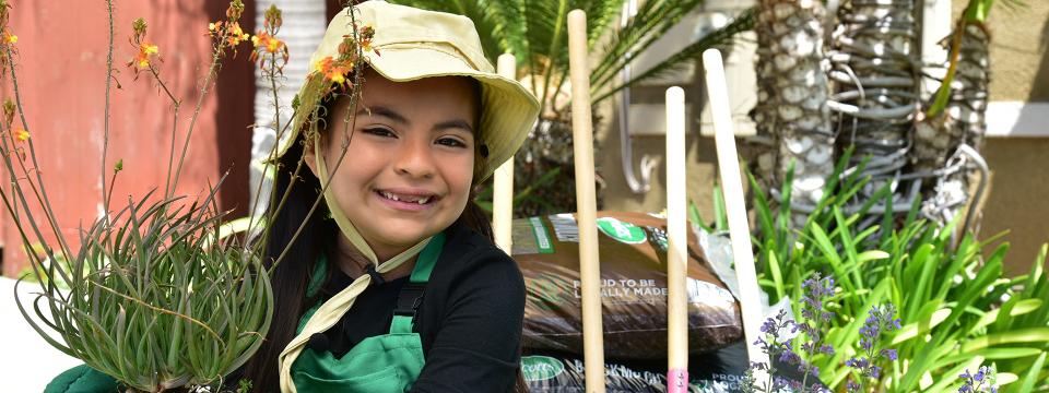 Child smiling wearing sunhat and gloves holding potted plant