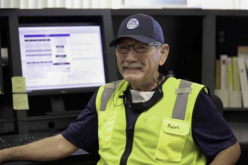 Zone 7 Employee smiling wearing a Zone 7 hat and safety vest with Ralph written on it, while sitting at a desk