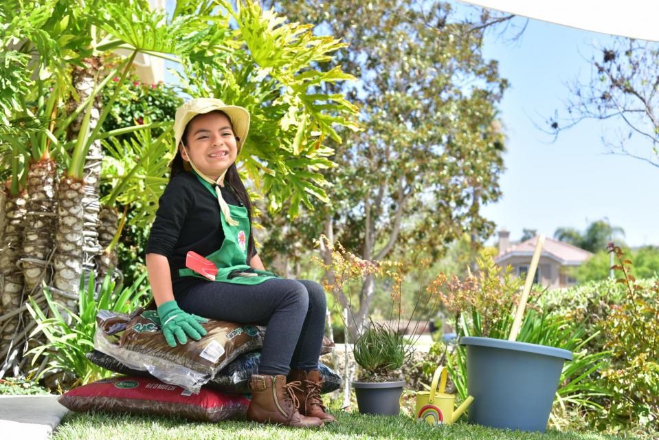 Water-Wise Wendy, a child smiling wearing a gardening hat, apron and gloves, sitting on bags of mulch in a garden